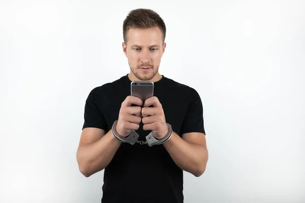 Beau jeune homme en t-shirt noir tenant smartphone tout en étant menotté sur fond blanc isolé — Photo