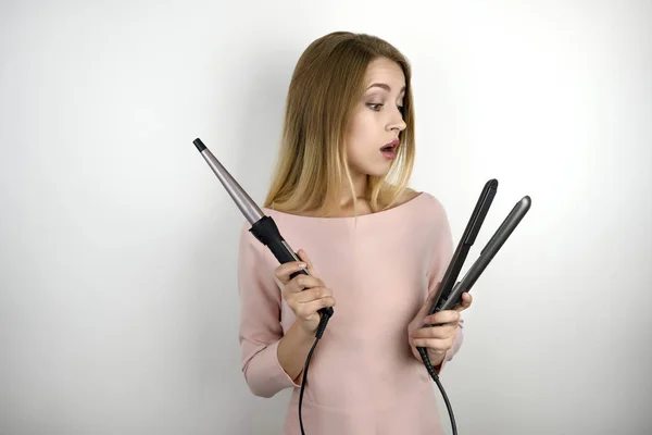 Joven hermosa mujer vistiendo vestido rosa de moda sosteniendo rizador y streightener pelo se siente feliz sobre fondo blanco aislado — Foto de Stock