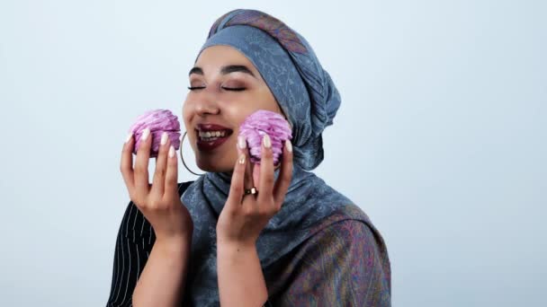 Joven hermosa mujer musulmana sonriendo sosteniendo sabrosos cupcakes en estado de ánimo juguetón sobre fondo blanco aislado — Vídeo de stock
