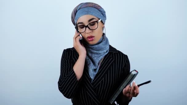 Fiatal gyönyörű Arab üzleti nő visel szemüvegek gazdaság tervező és toll birtoklás beszélgetés-val partner on smartphone-ra elszigetelt fehér háttér — Stock videók