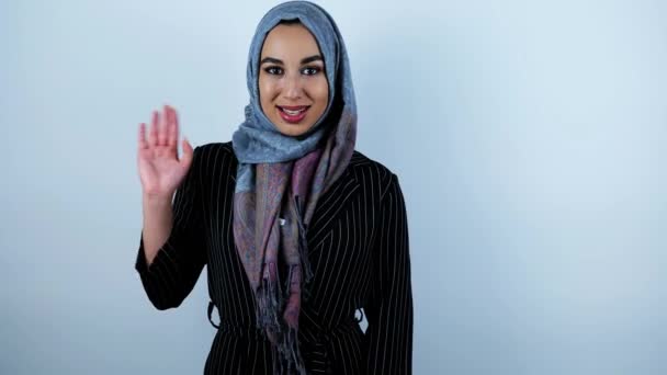 Wanita muslim muda yang cantik melambaikan tangannya dan dengan murah hati tersenyum pada latar belakang putih yang terisolasi — Stok Video