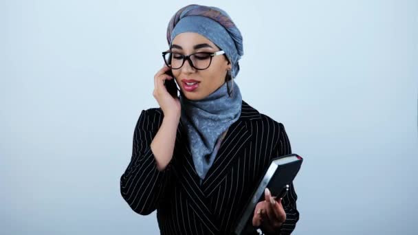 Wanita muda bisnis arab cantik mengenakan kacamata memegang perencana memiliki percakapan yang sukses di smartphone merasa bahagia di latar belakang putih terisolasi — Stok Video
