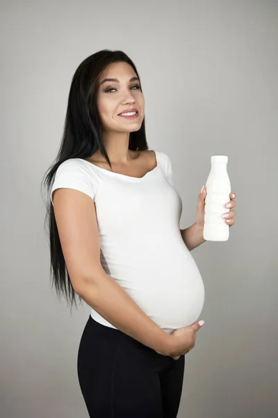 Beautiful pregnant brunette woman holding bottle with yogurt smiling on isolated white background — Stock Photo, Image