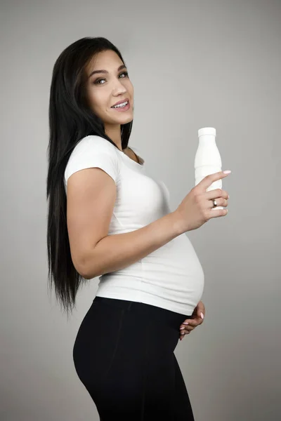 Bella donna bruna incinta in possesso di bottiglia con kefir sorridente in piedi mezzo giro su sfondo bianco isolato — Foto Stock