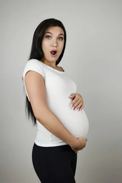 Mooie zwangere brunette vrouw houdt haar zwangere buik met verrassing op geïsoleerde witte achtergrond — Stockfoto