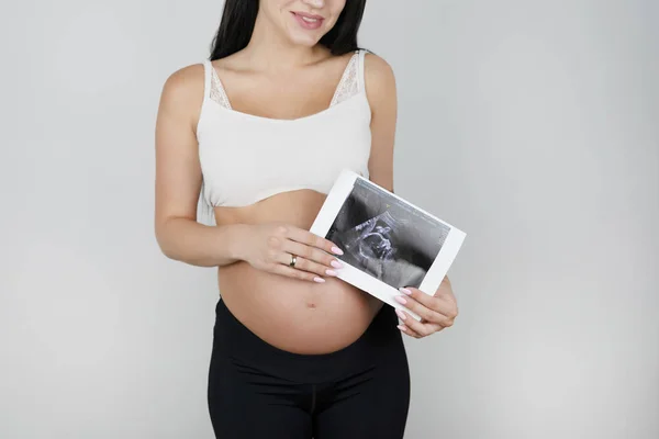 Pregnant brunette woman holding ultrasound picture of her baby on isolated white background — Stock Photo, Image