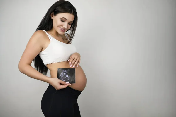 Bella donna bruna incinta che tiene l'immagine ad ultrasuoni del suo bambino su sfondo bianco isolato — Foto Stock