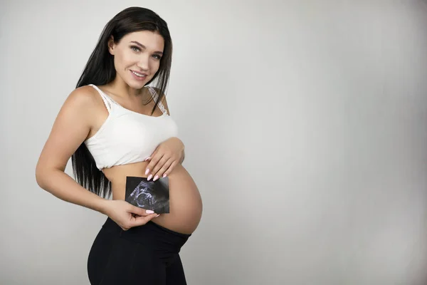 Bella donna bruna incinta che tiene l'immagine ad ultrasuoni del suo bambino loking felice su sfondo bianco isolato — Foto Stock