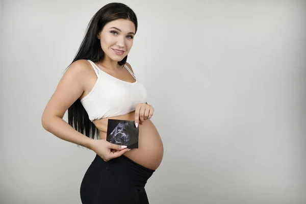 Bella donna bruna incinta che tiene l'immagine ad ultrasuoni del suo bambino che prova amore sorridendo su uno sfondo bianco isolato — Foto Stock