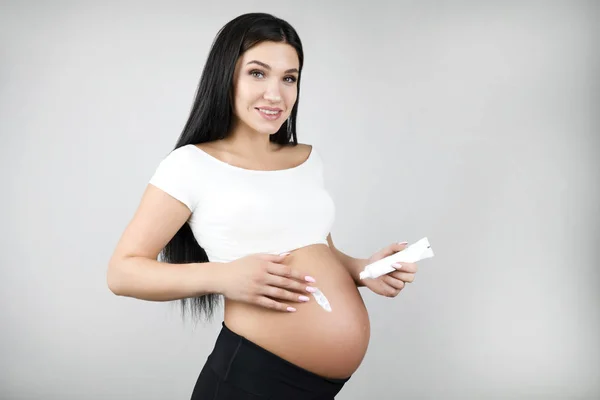 Gelukkig zwangere brunette vrouw het aanbrengen van Body Cream op haar buik staande een halve zet op geïsoleerde witte achtergrond — Stockfoto