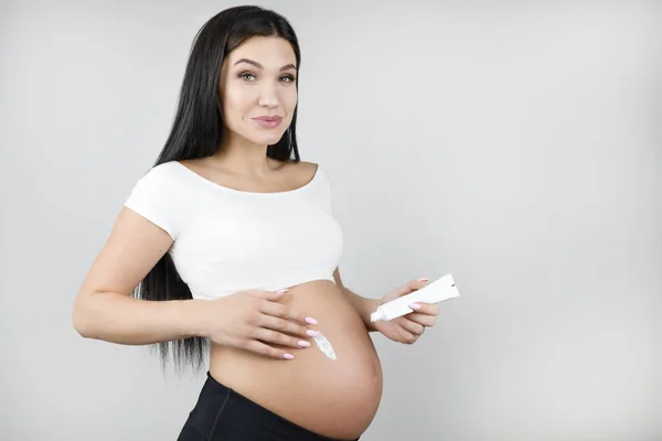 Gelukkige zwangere brunette vrouw die body lotion toepast op haar naakte buik staande een halve beurt op geïsoleerde witte achtergrond — Stockfoto