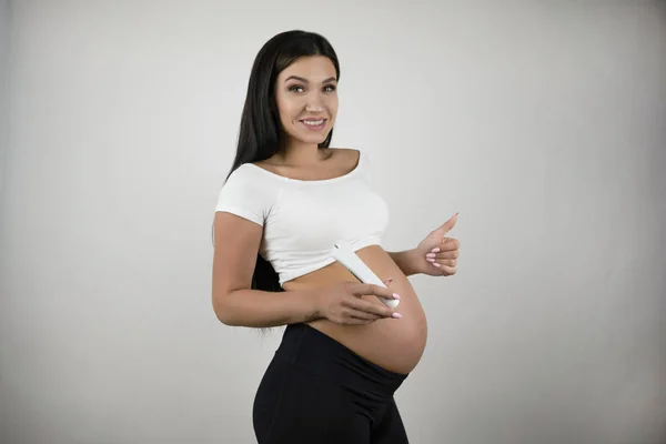 Giovane donna bruna incinta sorridente che applica burro corpo sulla sua pancia nuda in piedi mezzo giro su sfondo bianco isolato — Foto Stock