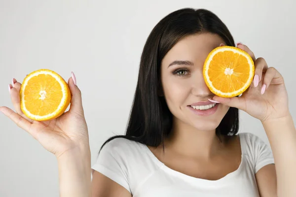 Jovem morena segurando uma metade de laranja perto do rosto e fechando o olho com outra metade no fundo branco isolado — Fotografia de Stock