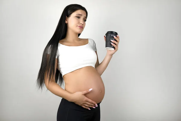 Bruna giovane donna incinta con tazza di caffè che dubita di berlo o non esita su uno sfondo bianco isolato — Foto Stock