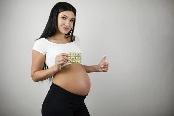 Bruna giovane donna incinta che tiene le vitamine in mano mostrando come segno su sfondo bianco isolato — Foto Stock