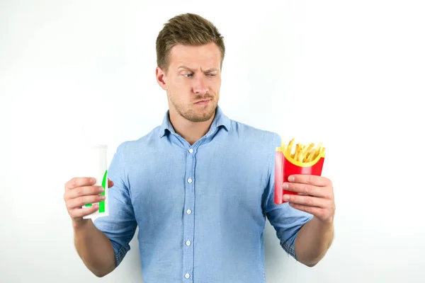 Knappe jonge man houdt soda in sanitair Cup en frietjes van fast food restaurant ziet er verdacht op geïsoleerde witte achtergrond — Stockfoto