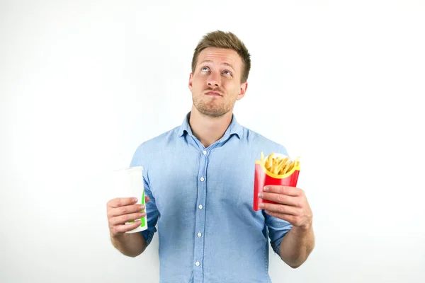 Knappe jonge man houdt soda in sanitair Cup en frietjes van fast food restaurant ziet er twijfelachtig op geïsoleerde witte achtergrond — Stockfoto
