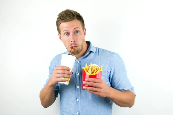 Knappe jonge man houdt soda in sanitair Cup en frietjes van fast food restaurant ziet er verbaasd over geïsoleerde witte achtergrond — Stockfoto