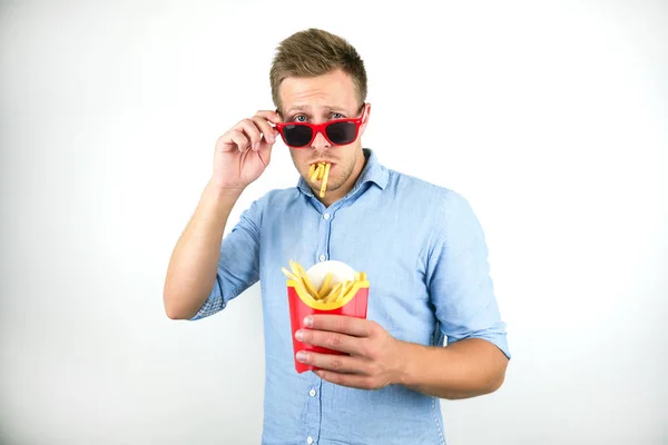 Grappige jonge man in rode brillen staan met frietjes in zijn mond op geïsoleerde witte achtergrond — Stockfoto