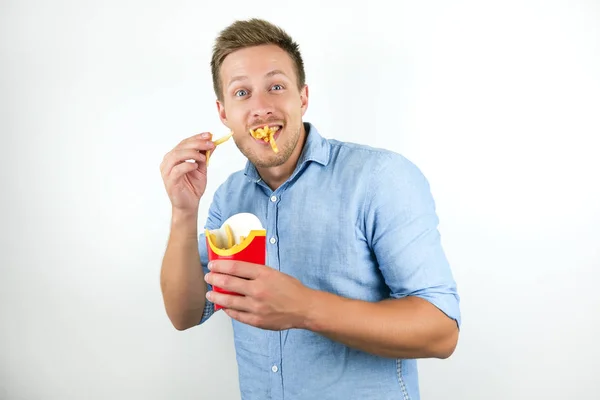 Jonge knappe man eet frietjes van fast food restaurant ziet er gelukkig uit op geïsoleerde witte achtergrond — Stockfoto