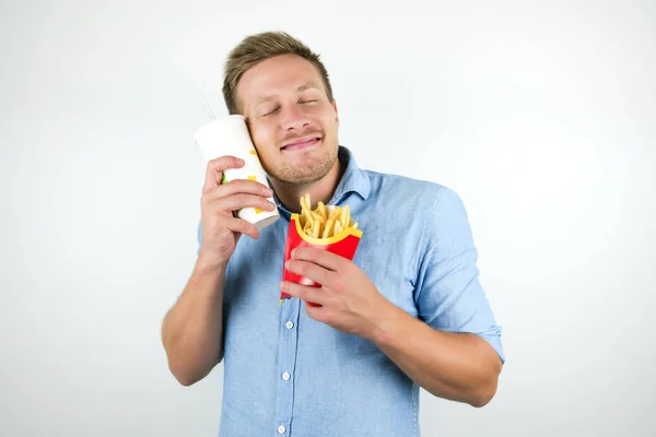 Jonge knappe man houdt papier beker met soda in de buurt van zijn tevreden gezicht terwijl eten eet frietjes van fast food restaurant op geïsoleerde witte achtergrond — Stockfoto