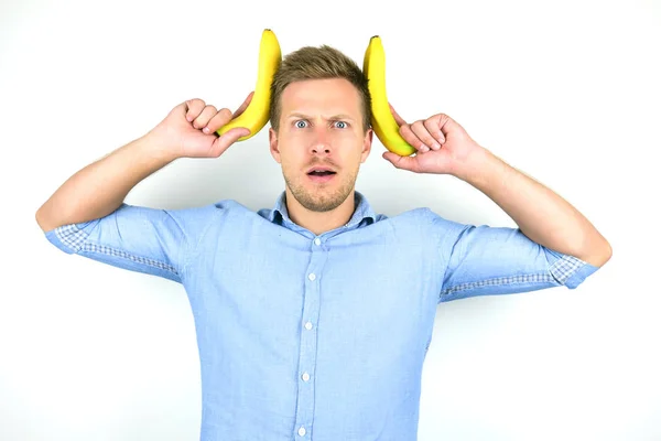 Jovem bonito homem segurando duas bananas frescas perto de sua cabeça olhando engraçado no isolado fundo branco — Fotografia de Stock