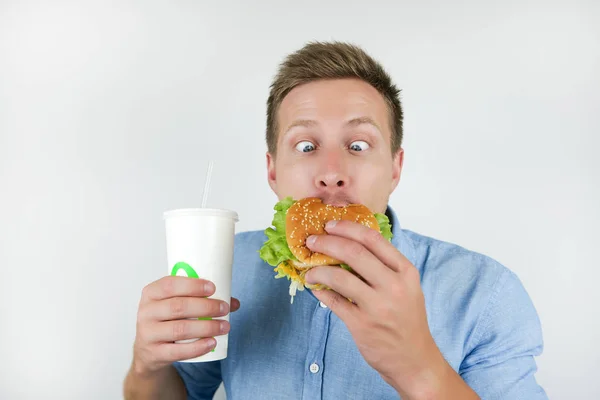 Young handsome man holding soda drink and biting burger from fast food restaurant looks funny on isolated white background — Stock Photo, Image