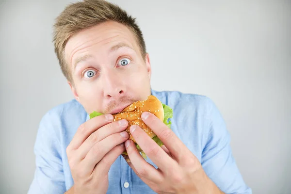 Pemuda tampan menggigit burger dari restoran cepat saji terlihat lapar pada latar belakang putih terisolasi — Stok Foto