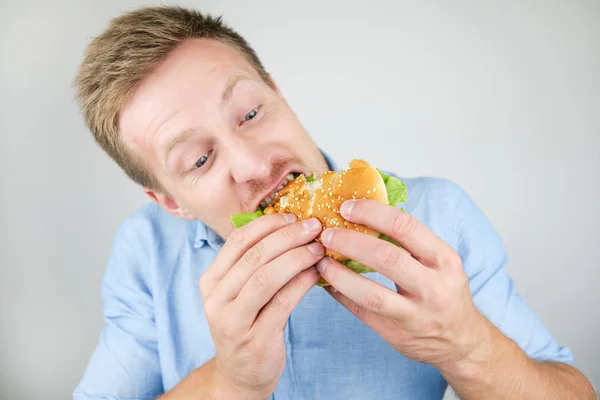 Jovem bonito homem mordendo saboroso hambúrguer de fast food restaurante parece com fome no fundo branco isolado — Fotografia de Stock