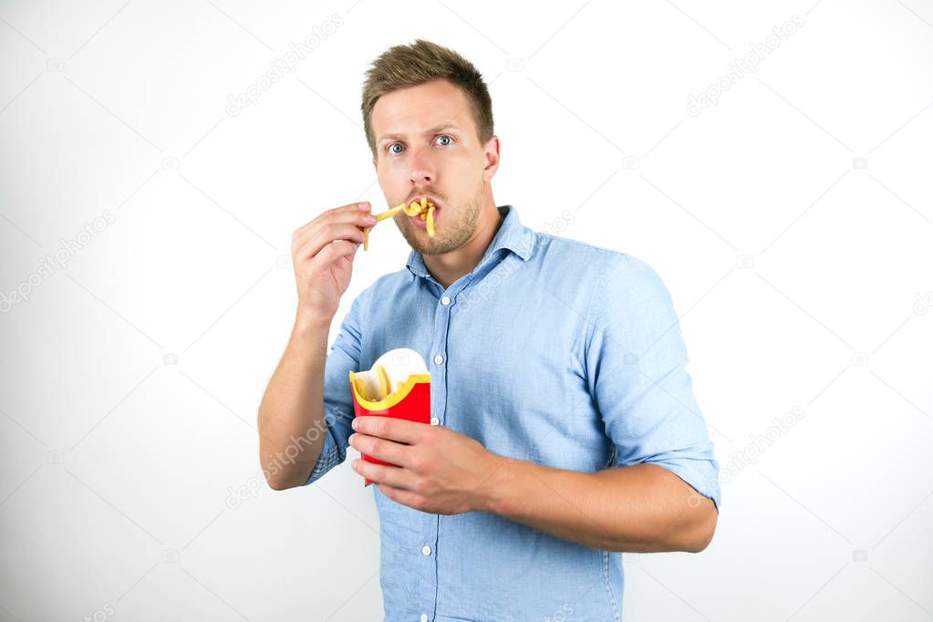 young handsome man eats french fries from fast food restaurant looks scared on isolated white background