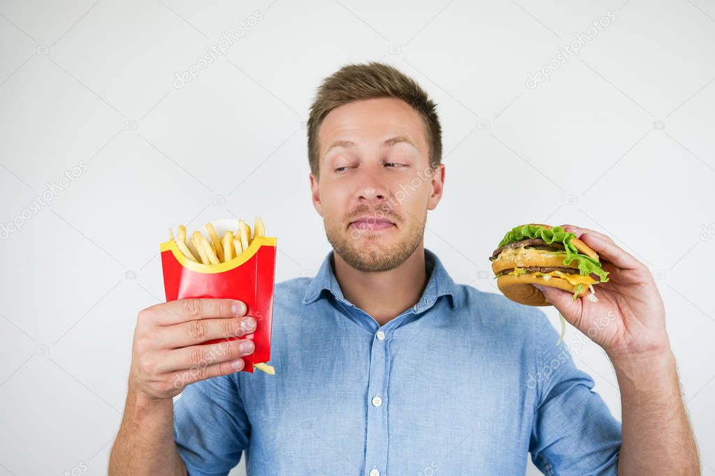 young handsome man holding fries and burger from fast food restaurant looks suspicious on isolated white background