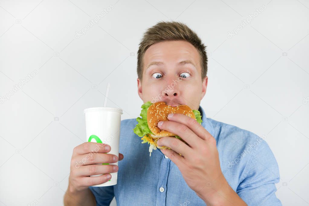 young handsome man holding soda drink and biting burger from fast food restaurant looks funny on isolated white background