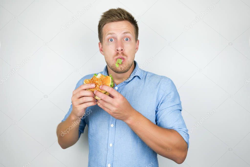 young handsome man biting cheeseburger from fast food restaurant standing with salad leaf in his mouth on isolated white background