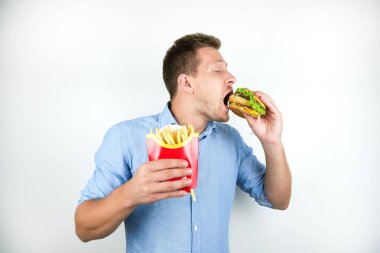 genç yakışıklı adam cheeseburger yiyor ve izole beyaz arka plan üzerinde fast food restorandan Fransız cips tutar