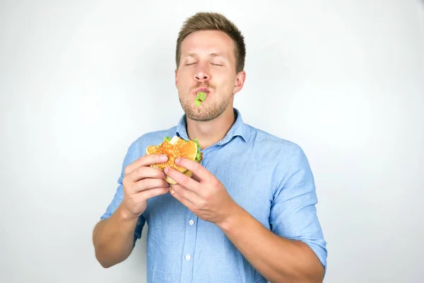Młody zabawny człowiek gryzący cheeseburger z Fast food restauracja stojący z liści sałaty w ustach na białym tle biały — Zdjęcie stockowe