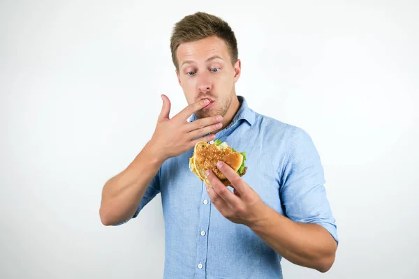 Jonge knappe man genieten van cheeseburger van fast food restaurant likken zijn vingers tijdens het eten op geïsoleerde witte achtergrond — Stockfoto