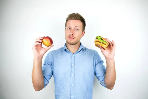 Jonge knappe man houdt Apple in de ene hand en cheeseburger van fast food restaurant in een andere op geïsoleerde witte achtergrond — Stockfoto