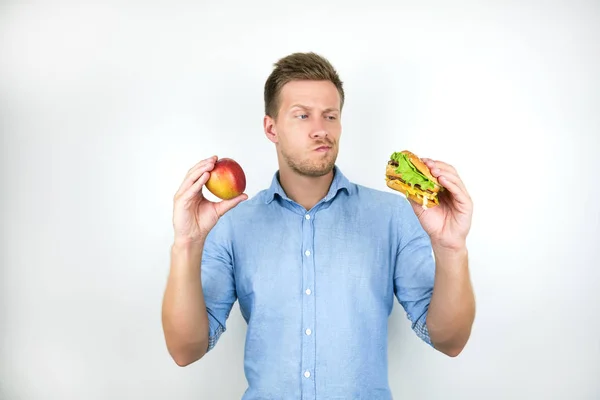 Jonge knappe man houdt Apple in de ene hand en cheeseburger van fast food restaurant in een ander gevoel twijfelachtig op geïsoleerde witte achtergrond — Stockfoto