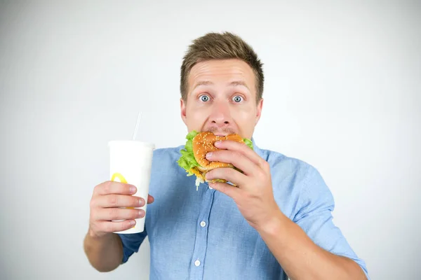 Jonge knappe man bijten cheeseburger van fast food restaurant en het houden van soda drankje gevoel hongerig op geïsoleerde witte achtergrond — Stockfoto
