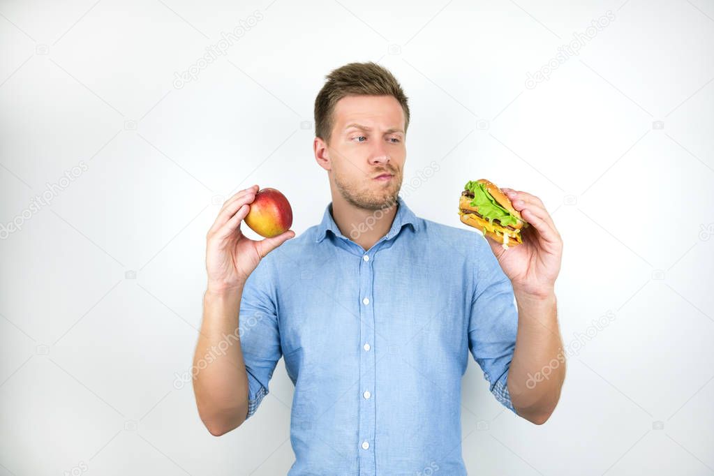 young handsome man holding apple in one hand and cheeseburger from fast food restaurant in another feeling doubtful on isolated white background