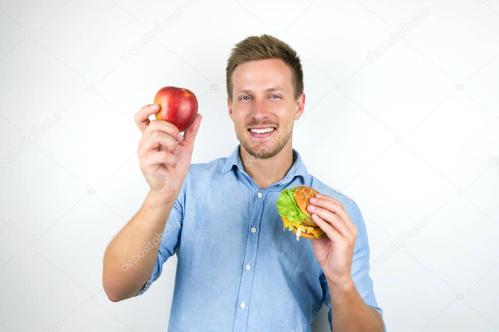 young handsome man holding apple in one hand and cheeseburger from fast food restaurant in another feeling happy on isolated white background