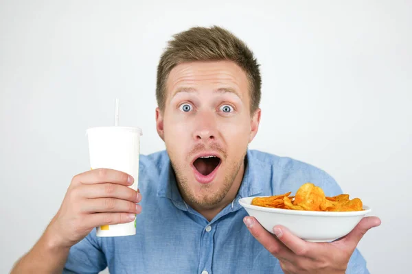 Jonge knappe man ziet er verbaasd uit terwijl hij soda drink en plaat met paprika chips in de buurt van zijn gezicht op geïsoleerde witte achtergrond houdt — Stockfoto