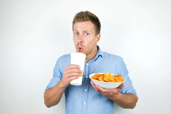 Young handsome man looks amazed while drinking soda and holding plate with paprika chips on isolated white background Stock Picture