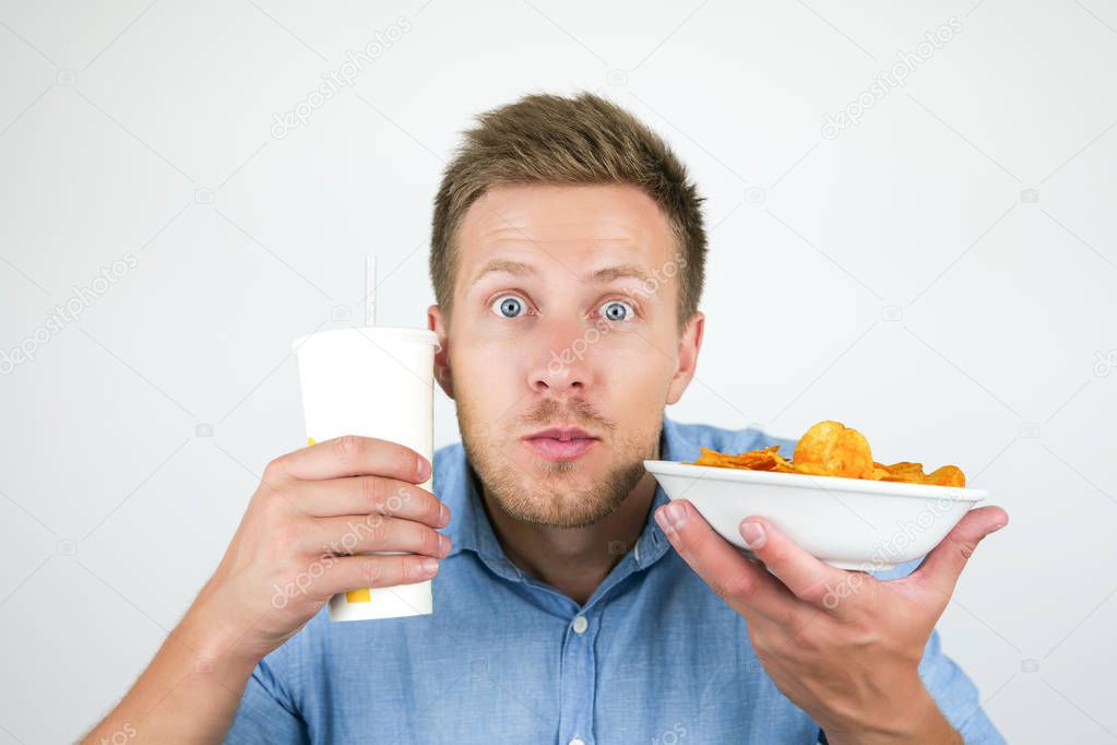 young handsome man looks amazed while holding soda drink and plate with paprika chips near his face on isolated white background