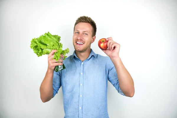 Jovem bonito segurando um monte de salada e maçã vermelha fresca sorrindo no fundo branco isolado — Fotografia de Stock
