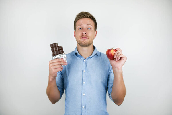 young handsome man holding chocolate bar in one hand and fresh ripe apple in another on isolated white background
