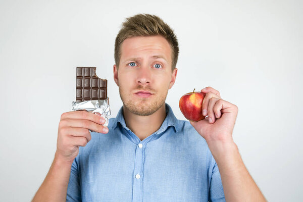 young handsome man holding chocolate bar in one hand and fresh ripe apple in another showing contrast on isolated white background