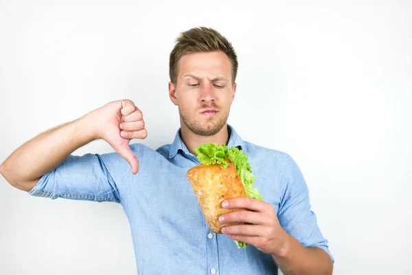 Jonge knappe hongerige man ziet er hongerig houden verse sandwich met salade blad tonen afkeer teken op geïsoleerde witte achtergrond — Stockfoto