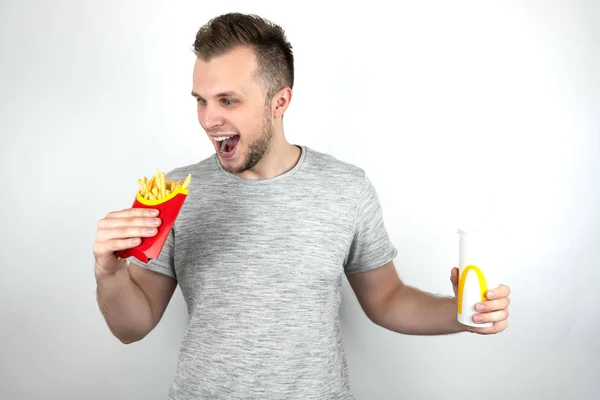 Jonge knappe man met frietjes en soda drink van fast food restaurant gevoel hongerig op geïsoleerde witte achtergrond — Stockfoto