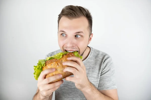 Jonge knappe hongerige man bijten verse sandwich met salade blad op geïsoleerde witte achtergrond — Stockfoto